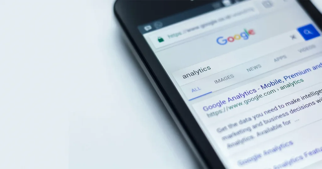 Smartphone exibindo uma busca no Google pelo termo 'analytics', com destaque para o link do Google Analytics, ferramenta usada para análise de dados e desempenho de sites e perfis online.