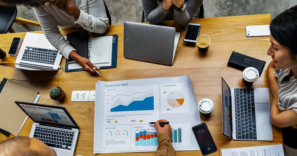 Equipe reunida em uma mesa de trabalho analisando gráficos de desempenho e estratégias de marketing digital com notebooks e relatórios visuais sobre crescimento de negócios.