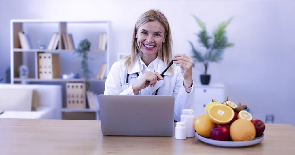 Nutricionista sorridente trabalhando em consultório com notebook, frutas e vegetais na mesa, simbolizando saúde, nutrição e atendimento online.